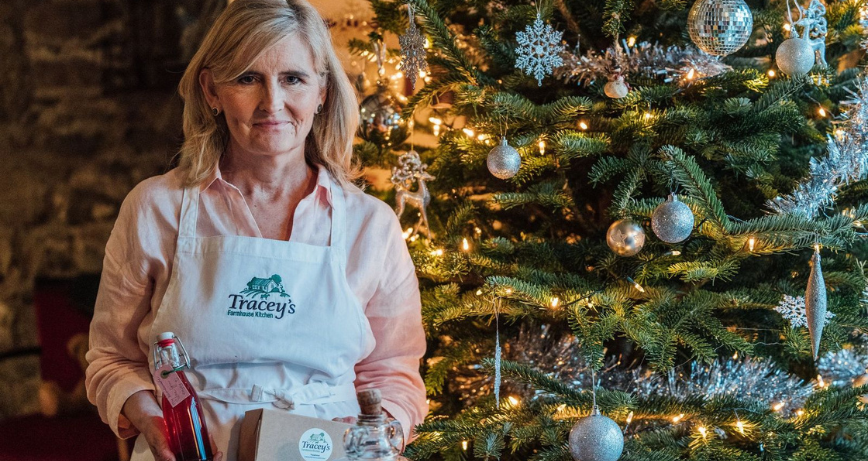 Tracey of Tracey's Farmhouse Kitchen standing beside a Christmas Tree holding foodie products